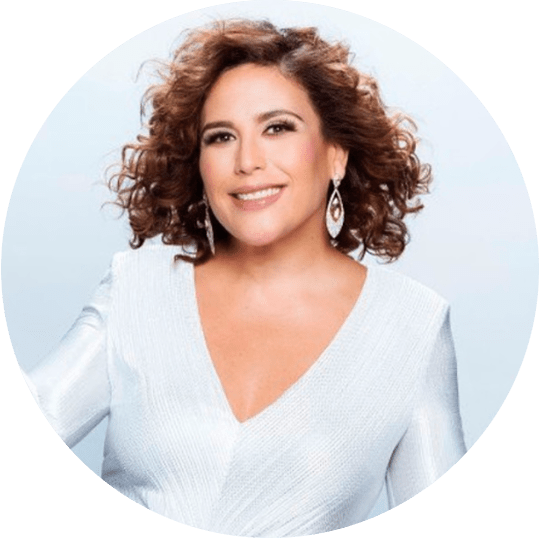 Angélica Vale smiling in a white dress with curly hair and earrings, posing against a light background.
