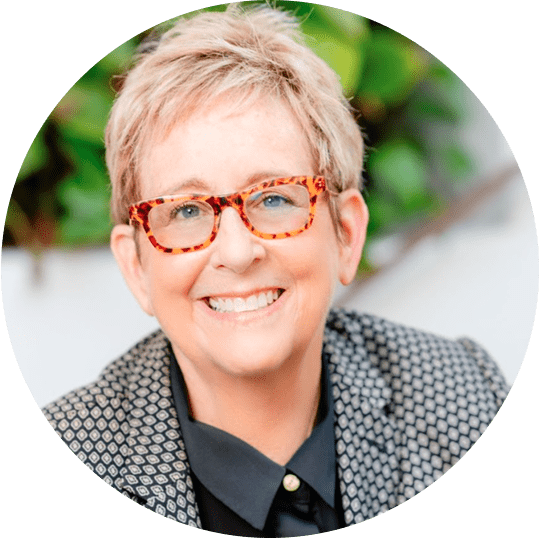Catherine Gray smiling at the camera, wearing tortoiseshell glasses and a patterned jacket, with greenery in the background.