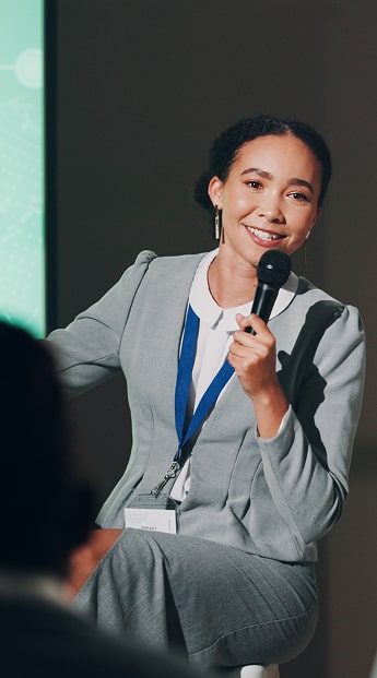Smiling woman in a gray suit holding a microphone, speaking at a conference or panel with a name badge and lanyard visible.