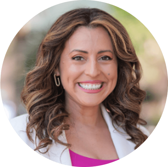 Ericka Aviles smiling with long, wavy hair, wearing a white blazer and pink top, and standing outdoors.