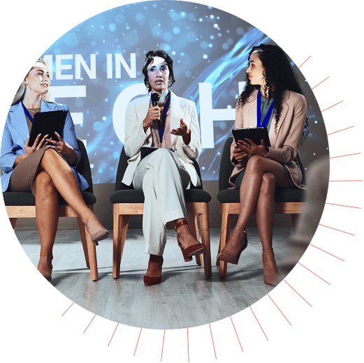 Three women seated on stage at a tech conference panel, with one speaking into a mic and others holding tablets.
